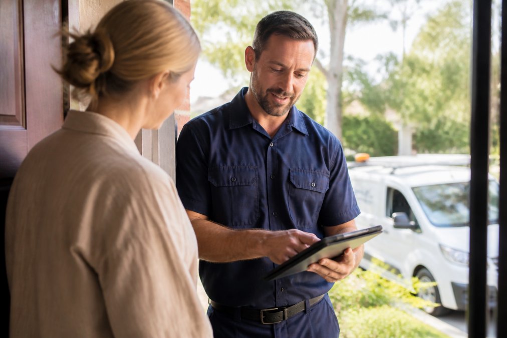 A professional plumber in a navy uniform holding a tablet and reviewing an itemised quote with a homeowner at their front door, bright morning light